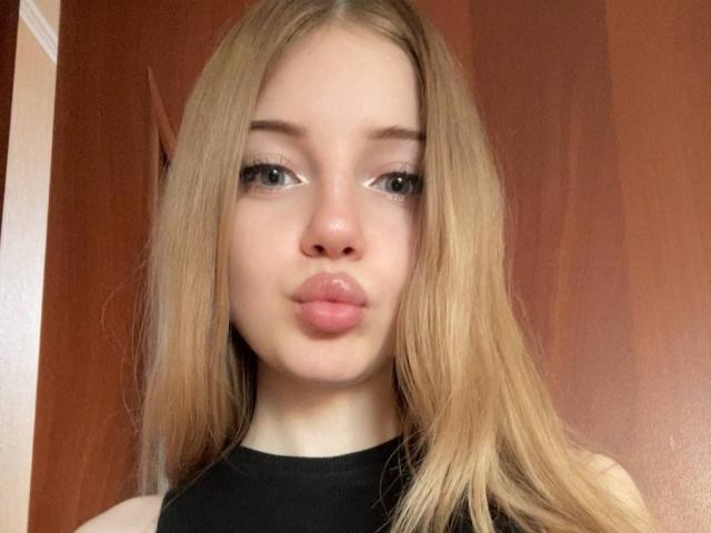 DandyMimi with blonde hair, pouty pink lips, and green eyes, wearing a black top, looking directly at the camera. Sheer, fair skin, slightly flushed, cute, and sexy.