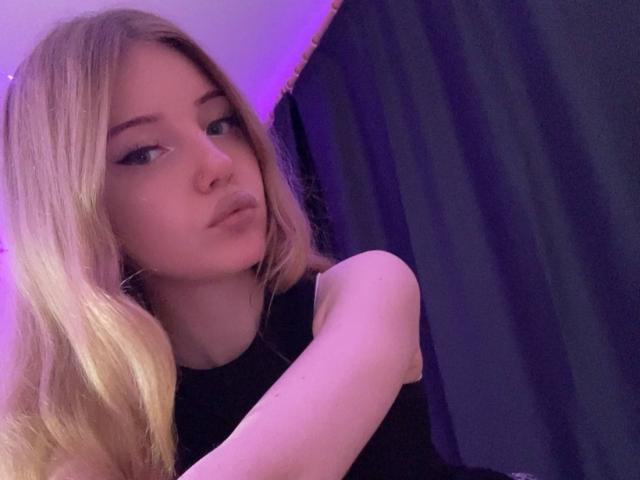 DandyMimi with blonde hair, pouty lips, and black sleeveless top, looking sideways with a slight smirk, purple lighting in background. Her ass is partially visible.