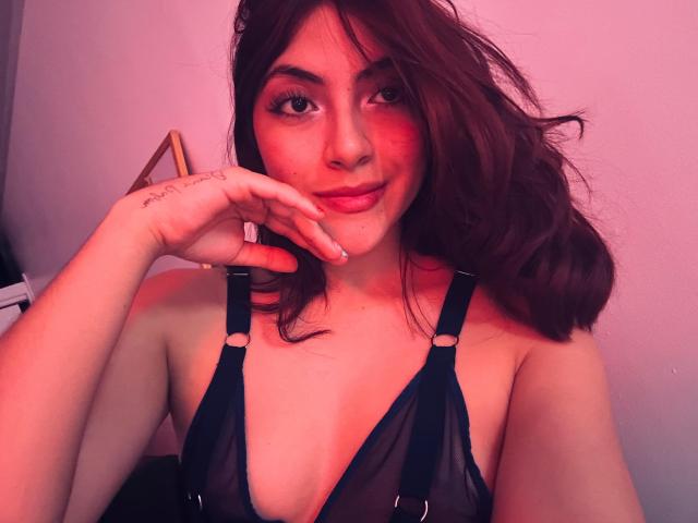 SpencerLe with wavy brown hair, pinkish-red lighting, black sheer lingerie, hand on chin, smiling, showing small tits, tattoo on finger, soft pink background.