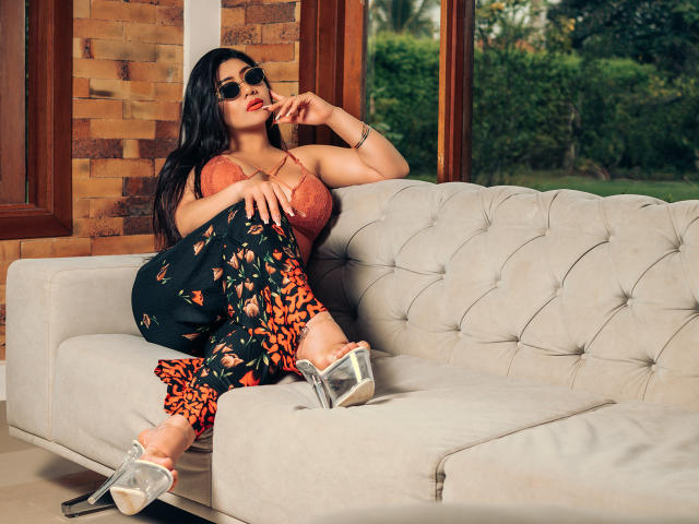 KhloeLawson with big ass, wearing orange lace top, black floral pants, silver heels, and sunglasses, lounging on a beige tufted sofa, finger near red lips, brick wall background.