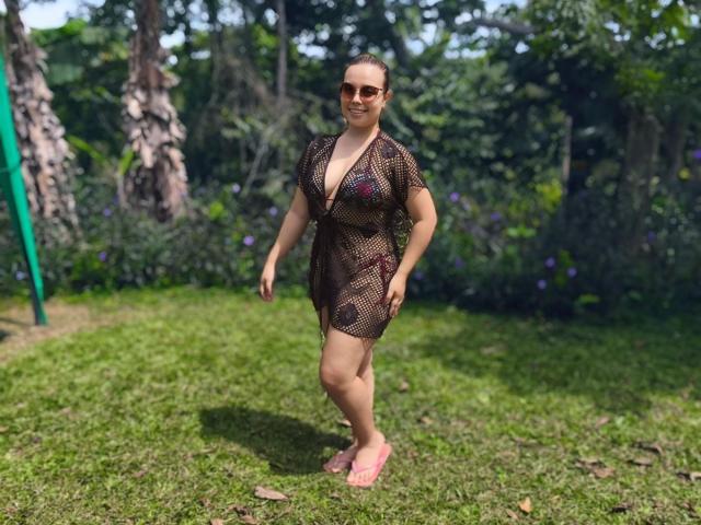 ZoeTobon69 wearing a see-through brown mesh dress, showing her tits, with pink flip-flops, standing on grass, sunglasses on, smiling, in a sunny forested area.