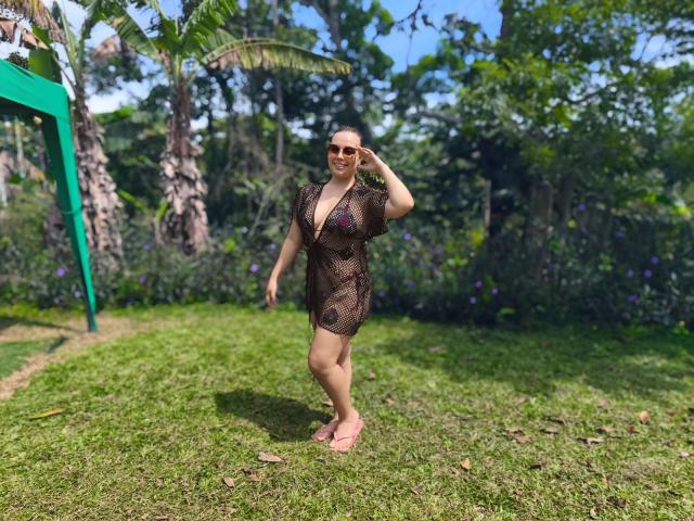 ZoeTobon69 with tan skin, brown mesh dress showing her tits and ass, pink sandals, sunglasses, standing on grass, hand in hair, tropical trees in background, sunny day.
