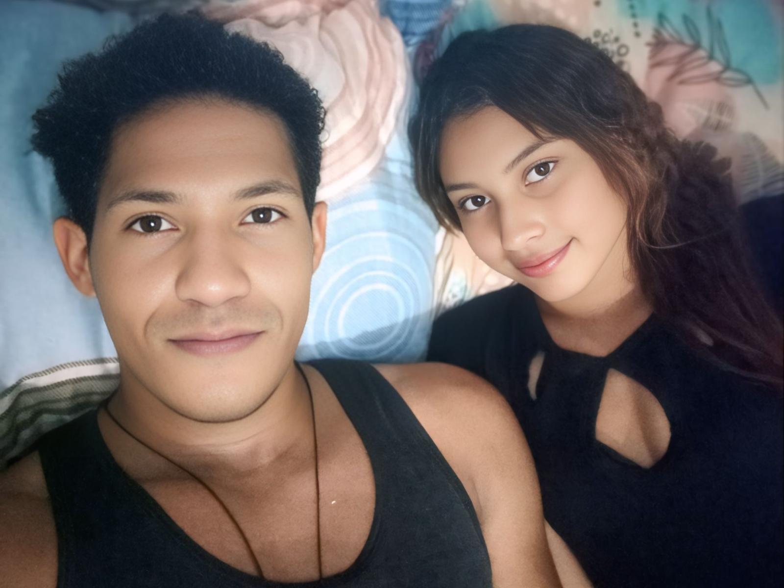 GibsonAndAsleys with short curly black hair, black tank top, and necklace, next to GibsonAndAsleys with long brown hair, black cutout top, smiling, on a bed.