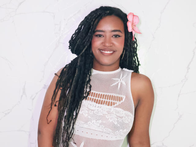 NaomiReyes with dark brown skin, long black braids, pink flower, white lace mesh top, smiling, showing her tits through the mesh, standing against a white wall.