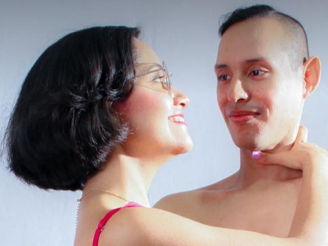 LizAndJuan with short black hair, glasses, and pink top, gently holds Juan's neck, who is shirtless with a shaved head, smiling at each other.