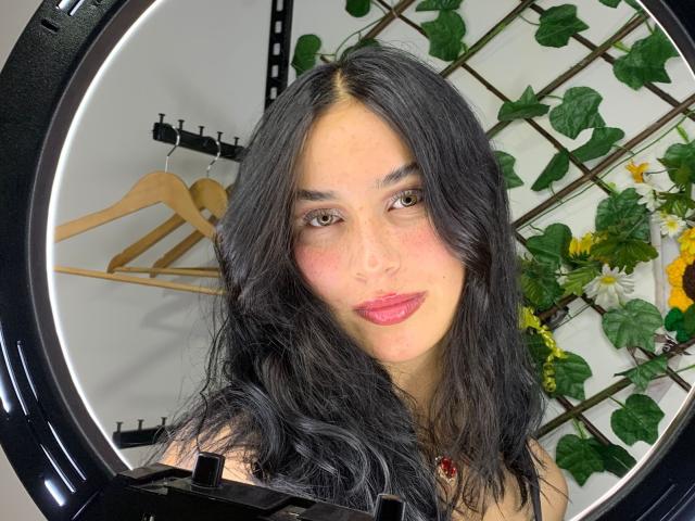 SolOrtega heeft lang, zwart, golvend haar, bruine ogen, roze lippenstift en een lichte blos. Ze kijkt ontspannen naar de camera en draagt een zwarte top..