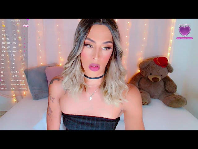 AmatistaBlue with blonde wavy hair, pink lipstick, black choker, and strapless black top, has a tattoo on her right arm, and a stuffed bear with a red hat.