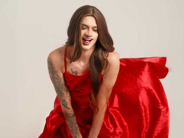 Elicarter with long brown hair, red satin dress, tattooed arm, and red pillow, smiling with mouth open, leaning forward. Boobs not visible, ass not visible, pussy not visible.