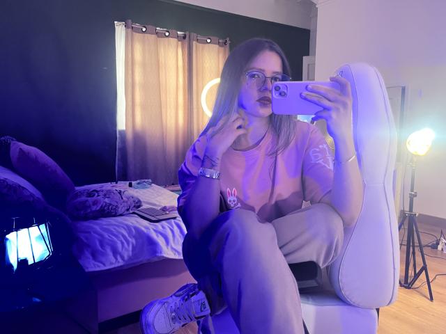 Alice, con gafas, camisa rosa y pantalones grises, sentada en una silla blanca, sosteniendo un teléfono, con zapatillas blancas, tomando una selfie con un espejo, iluminación azul y morada..