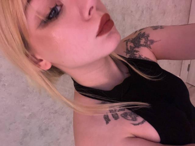 EnchantingEyes has blonde hair, dark lipstick, tattoos on her shoulder and chest, and is wearing a black halter top. Her blue eyes are slightly blurry in the photo.