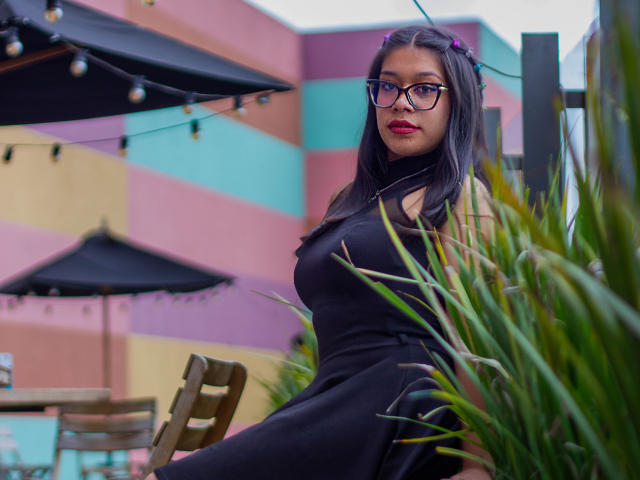 CuteVioletta with long dark hair, purple hairpins, glasses, and red lipstick, wearing a black dress, sitting in front of green plants, black umbrellas, and colorful building.