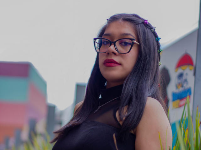 CuteVioletta with dark hair, glasses, red lipstick, and black mesh top, standing outdoors, looking slightly to the side, with colorful buildings blurred in the background.