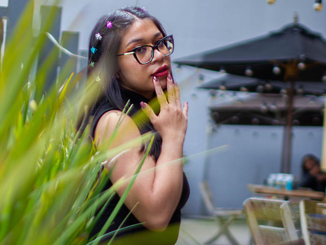 CuteVioletta with long black hair, wearing glasses, black top, and colorful hair clips, sucking on her finger, surrounded by green plants, looking cute and sexy.