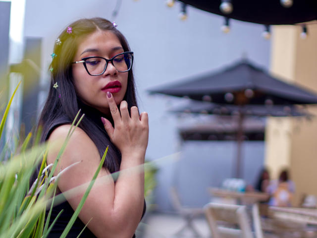 CuteVioletta with dark hair, glasses, pink flowers, red lipstick, finger to mouth, black sleeveless top, outdoor setting, blurred green plants, black umbrella, people in background, calm expression.