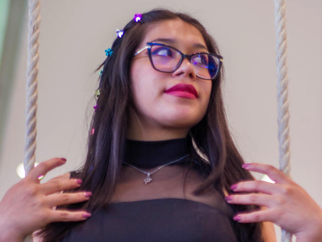 Violetta, com cabelos castanhos longos, uma tiara de flores, óculos azuis, batom rosa, uma blusa de malha preta, com as mãos no peito, dedos pintados de rosa, olhando para cima, seios visíveis, um colar, cordas brancas..