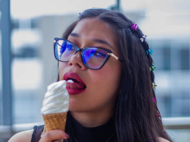 CuteVioletta with dark hair, wearing blue reflective glasses, a flower headband, and red lipstick, licking a vanilla ice cream cone. Her tongue is out, and she's holding the cone.