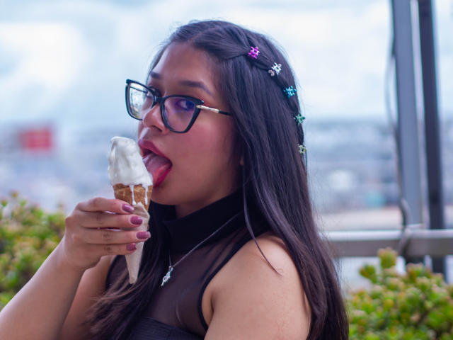 Violetta, com cabelos longos e escuros, óculos e unhas cor-de-rosa, lambendo um sorvete com cobertura de creme, vestindo uma blusa preta transparente, ao ar livre, com uma paisagem urbana desfocada ao fundo..