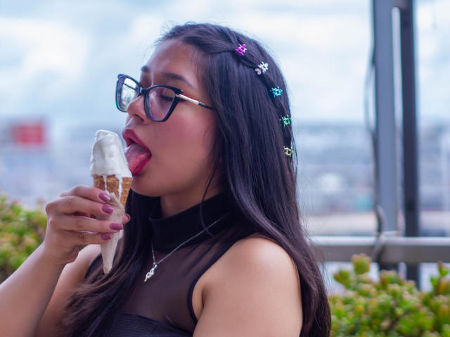 CuteVioletta with black hair, purple hairpins, and glasses, licking a white-frosted ice cream cone, wearing a sheer black top, showing pink lipstick and purple nails.