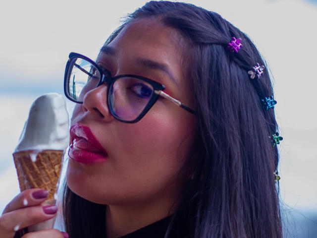 CuteVioletta with dark hair, pink flower hairpins, black glasses, red lipstick, holding a cone with a scoop of ice cream, looking up. Her ass is not visible.