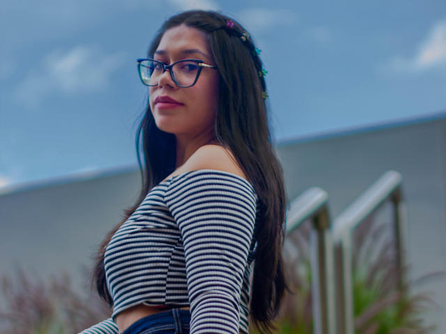 CuteVioletta with long dark hair, wearing glasses and a striped off-shoulder top, showing her big tits, looking over her shoulder, outdoors with a blue sky.