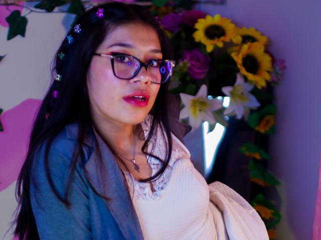 CuteVioletta with glasses, pink lipstick, and a white dress, wearing a blue cardigan, and flower hairpins, has a cute face and small tits, standing near a flower vase.