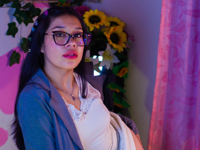 CuteVioletta with glasses, pink lipstick, white lace dress, blue cardigan, and flower headpiece, standing by sunflowers and pink curtains, looking at camera with a slight smile.