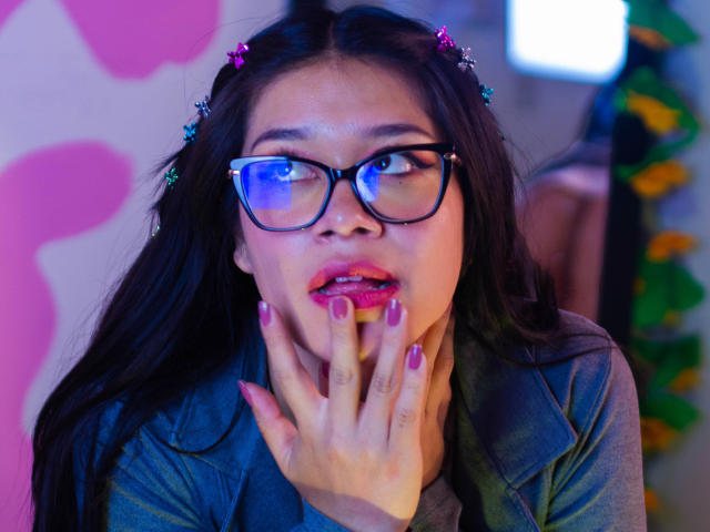 Violetta, com cabelo escuro, óculos e unhas rosa, tocando a boca com os dedos, vestindo uma jaqueta azul, tem grampos de cabelo coloridos e uma expressão ligeiramente surpresa..