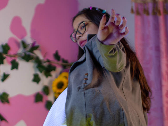 CuteVioletta with glasses, brown hair, and pink hairpins, reaches out with her right hand, wearing a gray button-up shirt, pink nail polish, and a white round object.