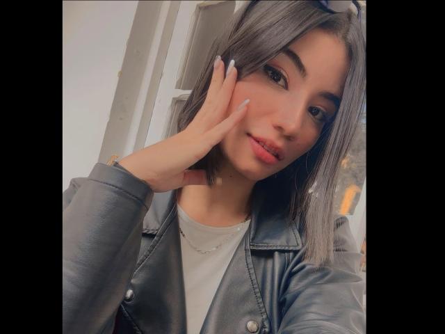 BerbyJohnson with medium tits, wearing a gray leather jacket, light gray shirt, and white nails, touching her cheek, brown eyes, light skin, and medium-length gray hair.
