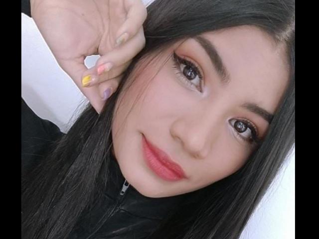 BerbyJohnson has long black hair, brown eyes, red lipstick, and is touching her ear with a yellow-nail-painted hand. She's wearing a black zip-up shirt.