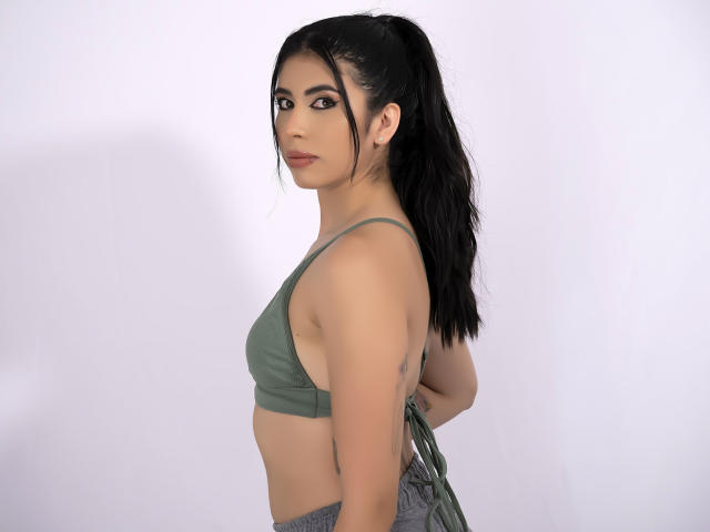 MiluSky with long black ponytail, green sports bra, showing small tits, tattoo on right arm, side profile, looking at camera, light skin, makeup, gray pants, white background.