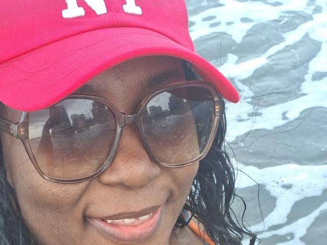 LucyLina69 lleva una gorra de béisbol roja, gafas de sol marrones grandes y sonríe con el pelo negro mojado, mostrando su piel marrón oscura y una camiseta naranja..