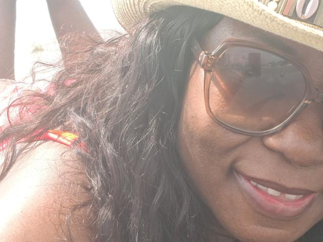 LucyLina69, con piel oscura, cabello negro ondulado, usando un sombrero de paja y grandes gafas de sol marrones, sonriendo, mostrando su top de bikini rojo y su lindo trasero a la luz del sol..