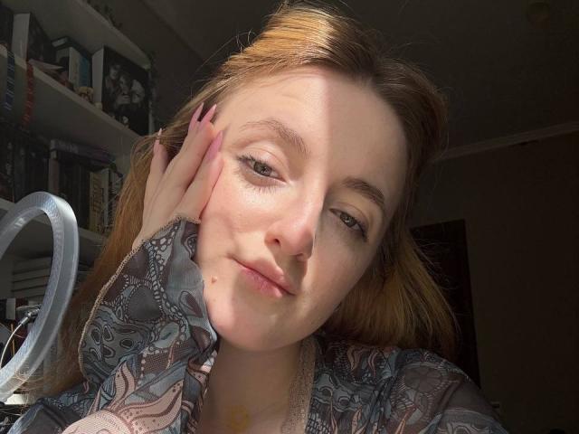 LunaBerryy with light brown hair, wearing a patterned shirt, hand on cheek, sunlight on face, pink nails, slightly smiling, looking at camera. Her tits are small and cute.