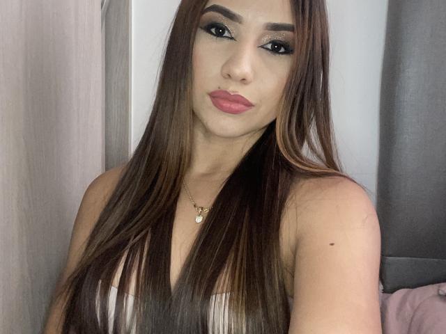 SofiaPeach with long brown hair, wearing a white top, has full pink lips, dark eyeliner, and a gold necklace. Her Tits are partially visible, showing a small mole.