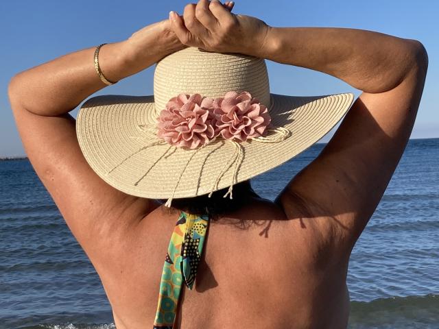 DeesseBleue's back is to the camera, wearing a straw hat with pink flowers, arms raised, showing sun-kissed skin and a colorful necktie, standing by the ocean.