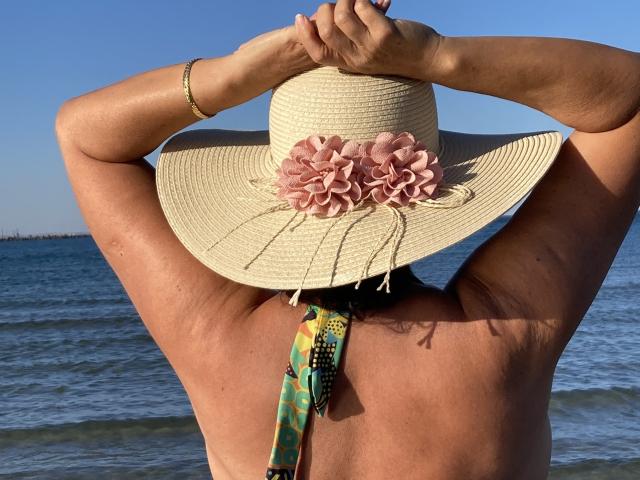 DeesseBleue's back, arms up, wearing a straw hat with pink flowers, no shirt, tan skin, yellow necklace, blue water, clear sky. DeesseBleue's tan arms and back are visible.