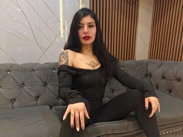 Mizuky, con lunghi capelli neri, rossetto rosso, top nero con spalline scoperte, pantaloni neri, seduta su un divano grigio imbottito, con un tatuaggio sulla spalla destra, un anello alla mano destra e le unghie dipinte..