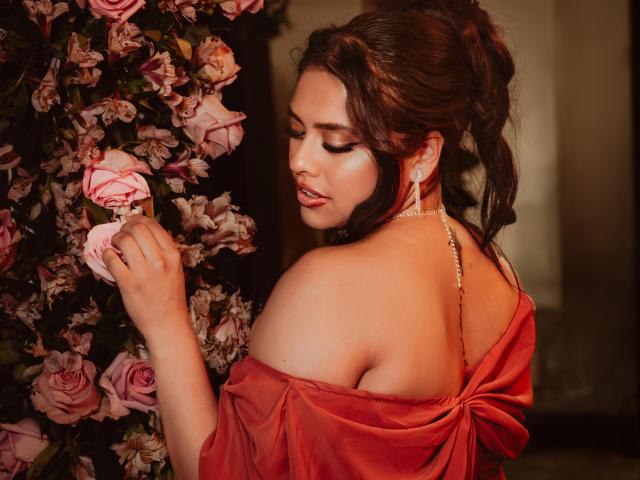 BellaSmithX69 with her back to the camera, wearing an off-shoulder red dress, holding pink roses, looking over her shoulder, showing her tits and a choker necklace.