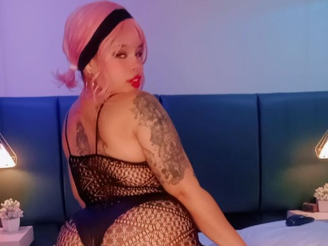 NatieRush with pink hair, black headband, red lipstick, black fishnet lingerie, tattooed arm, looking over shoulder, on bed, blue headboard, lamps, white wall, bedroom setting.