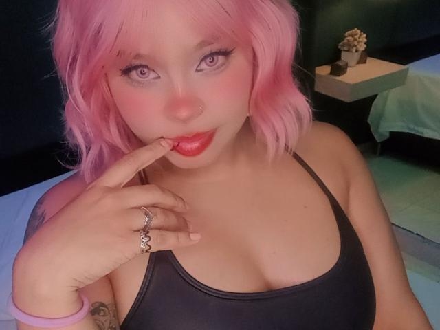 NatieRush with pink hair, purple eyes, red lipstick, black tank top, finger on lip, ring on finger, tattoo on wrist, looking at camera, cute, sexy.