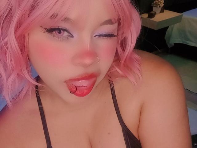 NatieRush with pink hair, winking, sticking out her tongue, nose ring, and red lipstick, showing off her tits in a black halter top, with purple eyeshadow.