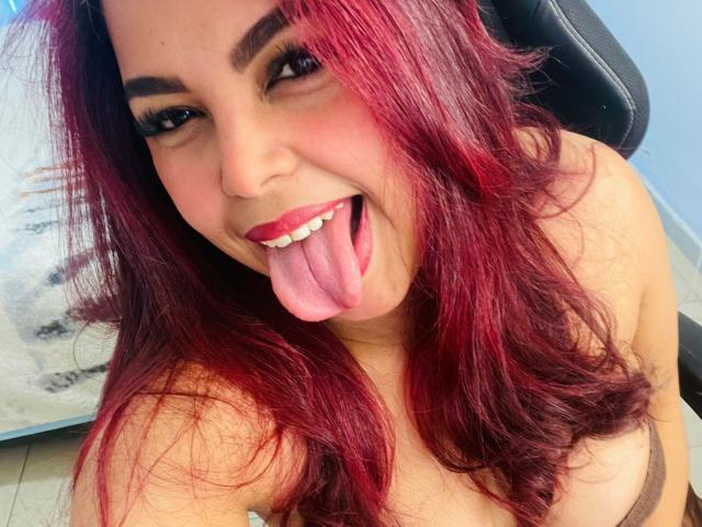 MorganMoretz with red hair, sticking out her tongue, showing white teeth, and wearing brown lingerie, has visible tits, and is smiling at the camera.