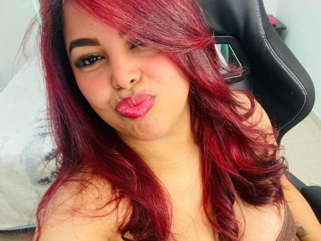 MorganMoretz with red and black hair, puffy pink lips, and thick eyebrows, winking and sticking out her tongue, showing off her small tits in a black chair.