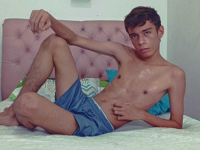 MaxSweett, shirtless, lying on bed with pink tufted headboard, wearing blue shorts, legs up, arm resting on knee, looking at camera, brown hair, slim, slightly sweaty.