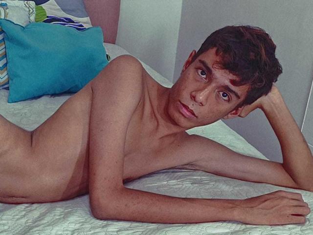 MaxSweett is a skinny, naked dude with short brown hair, lying on a bed, looking at the camera with a slightly curious expression, his arm resting on the bed.