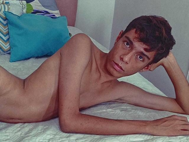 Naked MaxSweett, lying on a bed, with blue and green pillows, has brown curly hair, a slim build, and is staring at the camera with a serious expression.