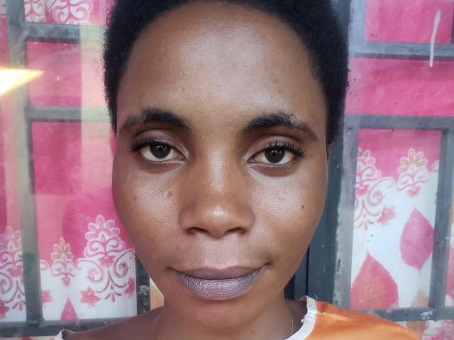 PaolaTtatts has dark brown skin, short curly black hair, and purple lipstick. Her eyes are medium brown with long eyelashes. She's wearing an orange garment.