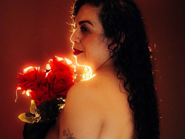 AlissaaMilf with curly black hair, red lipstick, and freckles, holding bright red roses, light glowing from behind, showing bare back and tattoo on left shoulder.
