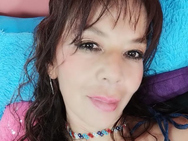 SamaraBellet's face, with pink lips, brown wavy hair, and dark eyeliner, wearing a colorful bead necklace, looking at the camera with a slight smile.